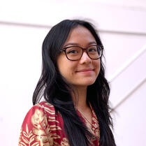 An Asian woman with long black hair and brown square glasses smiles slightly at the camera. She is wearing a red and tan button-down shirt.
